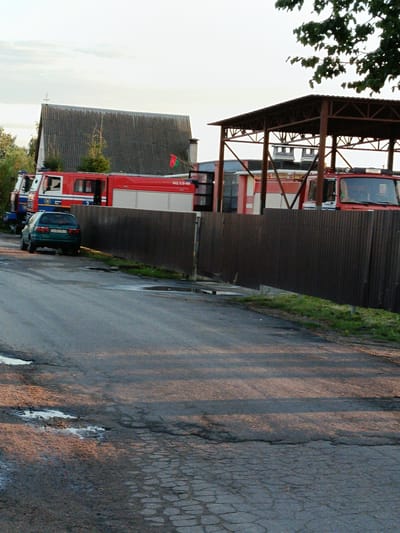 The image shows a rural scene with several fire trucks parked under a metal canopy. A dark green sedan is parked on the side of a cracked asphalt road in front of a corrugated metal fence. In the background, a building with a dark, sloped roof is visible, and trees line the property. The sky is overcast, suggesting it may be evening or a cloudy day. The location is identified as Smalyavichy, Smalyavichy District, Minsk Region, Belarus.