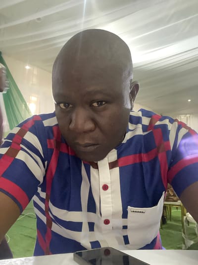 A man is photographed indoors, looking directly at the camera with a serious expression. He is wearing a short-sleeved shirt with a plaid pattern of blue, white, and red stripes. The shirt has a white collar and a placket with red buttons. A white pocket is visible on the right side of his chest, with a metallic tag that appears to say "DUFFY." The background consists of white draped fabric on the ceiling and walls, with some green fabric and lighting visible. The man's head and upper torso are the primary focus of the image. The scene is illuminated by overhead lighting, which creates some glare on the ceiling. The man is positioned in the foreground, with the background out of focus. The image suggests an indoor event or gathering, possibly a formal occasion, given the draped fabric. The man appears to be of African descent, with dark skin and a bald head. The image does not contain information that definitively places the location in Unguwar Sarki, Nigeria.