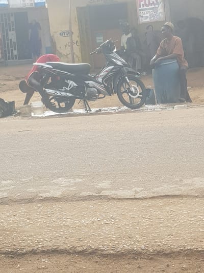 The scene is an exterior shot of a street in Tukur Tukur, Nigeria. A black and silver motorcycle is parked on the right side of the street. A man in a red shirt is crouched down near the motorcycle. Nearby is another man in a tan shirt and blue clothing is visible near a building. There is a group of people in the background near a building with a sign that says "SERVICE". The street is paved.