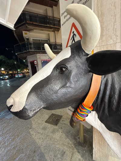 The image shows a close-up, low-angle shot of a large, black and white cow sculpture. The sculpture has a prominent white horn and a leather collar around its neck. The collar is decorated with a rainbow-colored ribbon, suggesting a pride theme. The cow sculpture is positioned outdoors, with a textured concrete ground and a building visible in the background. A warning sign with a pedestrian symbol is partially visible beneath the cow's head. The building appears to be made of brick and has several windows. Streetlights and some trees are visible in the distance, indicating it might be evening or night. The overall lighting suggests artificial light sources are present, possibly from streetlights or shop fronts. There is some visible text on a sign beneath the cow's head, though it is largely obscured and unreadable. The scene appears to be in an urban or town setting.