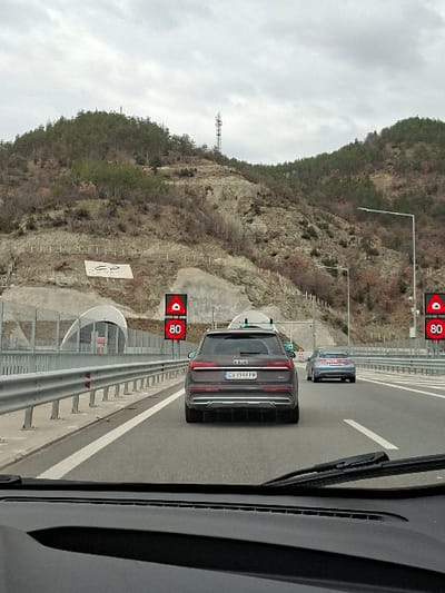 This image depicts a multi-lane highway in Jeleznitsa, Bulgaria, viewed from inside a vehicle. The dashboard and windshield wipers are visible in the lower foreground. A dark brown Audi Q8 SUV, bearing Bulgarian license plate 'CB 9944 PM' and equipped with roof-mounted sensors, is centrally positioned in front of the camera vehicle. Ahead of the Audi, a silver sedan proceeds in the same lane. The highway features metal guardrails to the left. Two red digital overhead signs display a warning triangle symbol and the numeral '80', indicating a speed restriction. Infrastructure resembling tunnel entrances or overpasses is visible on the left side of the highway. The surrounding landscape consists of rugged hillsides with sparse evergreen and deciduous trees, exposed rock, and soil. A tall communication tower is present on a distant hill to the left, and a large white "СР" inscription is visible on the lower left hillside. The sky is uniformly overcast.