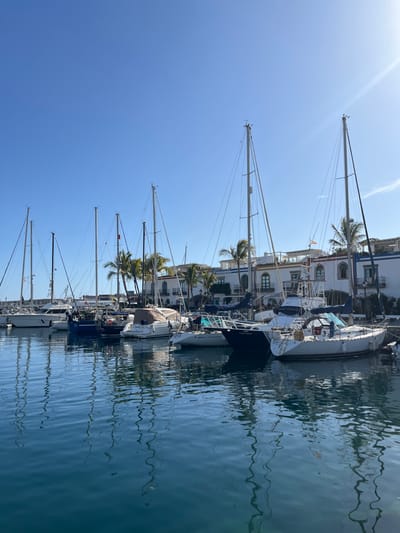 The image shows a marina in Puerto de Mogán, Gran Canaria, Spain. The coordinates (latitude: 27.817, longitude: -15.766) are located near the marina. The image shows a clear, sunny day. The marina is filled with sailboats and yachts. There are buildings behind the marina, which are all white, with flat roofs and some palm trees in front. The marina is calm and there are no people visible in the image. A few of the sailboats are docked and others are floating freely. The boats are all white and blue. The water is clear and blue and the sky is clear and blue. There are no other objects or people in the image. There is a lot of reflection in the water.