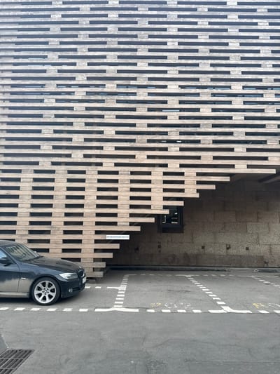 A black BMW sedan is parked in a marked parking spot in front of a modern building with a facade of horizontal wooden slats. The building has a recessed entrance area with a granite-tiled wall and a small window. A sign on the wooden facade reads "НЕГАБАРИТНОЕ МЕСТО," which translates to "Oversized Parking." The parking lot is paved and shows painted white dashed lines delineating parking spaces. A metal grate is visible on the left side of the frame. The scene is outdoors during daylight.