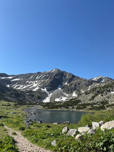 The image depicts a serene mountain landscape with a clear blue sky. In the foreground, a dirt path winds through lush green grass and scattered rocks. A calm lake with dark, reflective water occupies the midground, its edges lined with more rocks and vegetation. Behind the lake, a majestic mountain range rises, its slopes covered with patches of snow and rugged grey rock formations. Sparse green shrubs dot the lower slopes of the mountains. The scene appears to be a natural, undeveloped area, likely a tourist destination in or near the city of Samokov, Bulgaria, given the context. There are no people or animals visible in the image, and the overall atmosphere is tranquil and natural.