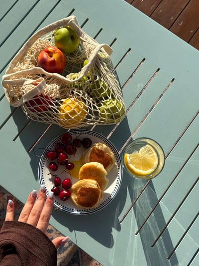 The image depicts a flat lay of a breakfast spread on a light blue slatted outdoor table, bathed in warm sunlight suggesting late afternoon or early morning. To the left, a cream-colored mesh bag contains several green apples and a yellow lemon. A single peach, with red and yellow skin, is also visible in the bag.
To the right, a white plate with a blue rim holds three small, golden-brown pancakes and a generous serving of dark red cherries, some still attached to their stems. A clear glass filled with water and a slice of lemon sits above the plate. The sunlight casts distinct shadows across the table, highlighting the texture of the wood.
In the bottom right corner, a person's hand with white nail polish adorned with small white polka dots is reaching towards the plate of food. The setting appears to be an outdoor patio or deck, with a glimpse of a wooden floor and a patterned rug in the background. The overall mood is relaxed and inviting, suggesting a leisurely meal enjoyed outdoors. There is no visible text in the image.