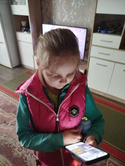 A young girl with blonde hair, wearing a pink vest over a green long-sleeved shirt, is looking down at a smartphone in her hands. She is standing in what appears to be a living room, with a television in the background and furniture, including a tall cabinet, visible. A rug is on the floor. The image is taken from a slightly elevated angle. The girl's expression is focused on the screen. The environment suggests a domestic setting within the Крывасельскі сельскі Савет, Belarus.