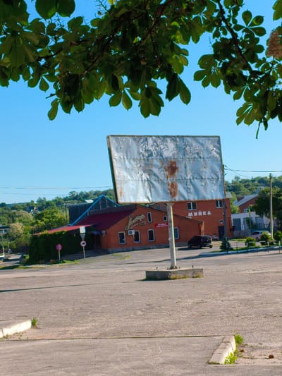 The image captures a roadside scene in Velyka Bahachka, Ukraine, under a clear, bright blue sky, suggesting daytime, likely morning or early afternoon. The foreground is dominated by a large, empty asphalt parking lot or open space, with a concrete curb in the bottom right corner. Overhanging green leaves from a tree, possibly a chestnut, frame the top of the photograph.

A prominent feature in the midground is a large, rectangular billboard made of corrugated metal, showing considerable weathering, rust, and peeling paint. It stands on a single concrete pillar in the center of the frame, currently advertising nothing. Behind the billboard are two brick buildings with red roofs. The building on the left, which appears to be a cafe or shop, has a sign displaying "Джерело" (Djerelo) and smaller text below that reads "ПРОДАМ" (FOR SALE), a phone number "(050) 933 93 69", and "ПРОДАЖ" (SALE). The building to its right has the word "МИЙКА" (CAR WASH) prominently displayed on its facade. A dark-colored car is parked between these two buildings.

Several small road signs are visible near the left building: a red circular sign with a white horizontal bar (indicating 'no entry' or 'forbidden') and another circular sign with a red border and a '0' inside, possibly indicating a parking restriction or local regulation. The background features a hilly landscape covered with lush green trees and scattered houses, indicating a semi-rural setting. The overall impression is one of a functional, albeit slightly rundown, commercial area in a small town.