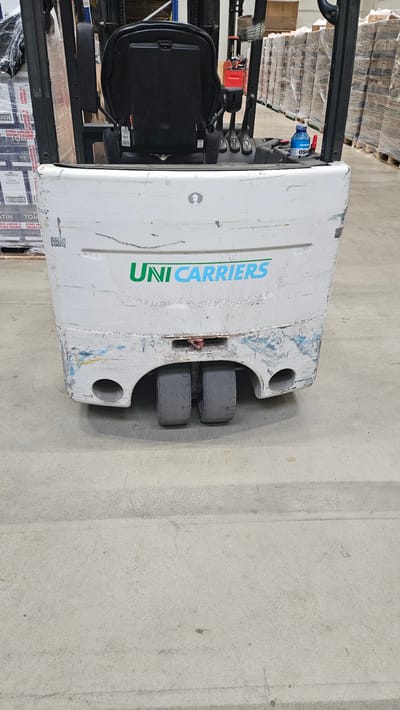 The image shows the front end of a white forklift. The forklift has "UNI CARRIERS" written in green and blue on its front. It has two large wheels at the bottom front. There are some scuff marks and minor damage on the white exterior of the forklift. To the left and behind the forklift, stacks of boxes wrapped in plastic are visible. In the background, there are more stacks of palletized goods. To the right of the forklift, there is a blue bottle and a red object. The forklift is on a concrete floor. The scene appears to be in a warehouse or storage facility. This scene is not located in the city ofĶekava, Latvia.