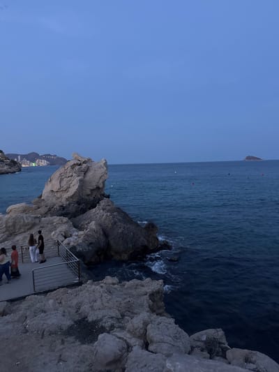 The image depicts a twilight scene at a rocky coastline in La Cala, Spain. The sky is a deep blue, transitioning from dusk to night. A large, weathered rock formation dominates the left side of the frame, jutting out into the dark blue ocean. A paved walkway with a metal railing runs along the edge of the rocks, where a small group of people are visible. Two individuals, a man and a woman, stand near the railing, looking out at the sea. To their left, two other people, possibly a child and an adult, are walking along the path. In the distance, the lights of a coastal city gleam across the water. Several buoys are scattered across the sea, and a small island is visible on the horizon. The waves gently lap against the rocky shore, creating white foam.