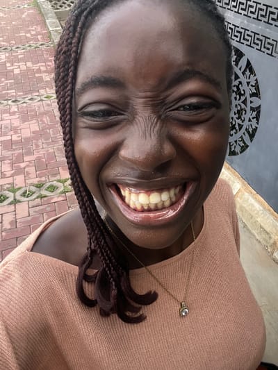 A close-up, frontal view of a young Black woman's face, captured from the shoulders up, reveals her broad, toothy smile. Her eyes are crinkled in mirth, and her expression is lively. She has dark, braided hair that falls over her left shoulder, and she is wearing a light peach-colored knitted top with a delicate gold necklace featuring a small pendant. The background shows a textured, grey wall with a geometric design and a section of red brick paving, suggesting an outdoor urban setting. The lighting indicates it is daytime. While the image doesn't explicitly state the location as Karum, Nigeria, the context implies an urban environment within the country.