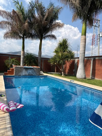 The image shows a swimming pool in an outdoor setting. The pool is tiled in blue mosaic and has a striped inflatable pool toy near the edge. Reflections of palm trees and the sky are visible on the pool surface. Tall palm trees stand near the pool. A brick building is adjacent to the pool area. A power transmission tower is visible in the background. Mountains are visible in the distance. The image was taken in San Francisco Lachigoló, Mexico.