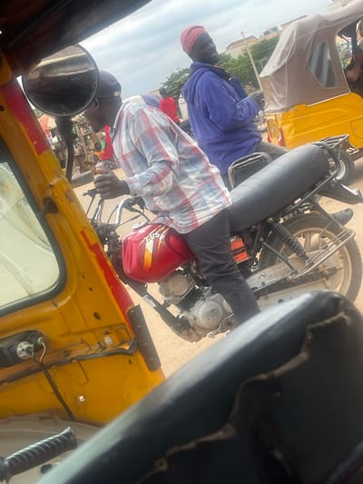 The image depicts a street scene in Galadimawa Village, Nigeria. Three people are visible, two men and a woman. One man is driving a yellow motorized tricycle, also known as an "Okada" or "Keke Napep." He is wearing a red and white checkered shirt and a dark head covering. Seated behind him is a woman wearing a purple jacket and a red headscarf. Another man, possibly a passenger, is visible in the foreground, wearing a dark head covering and a red and white checkered shirt. The tricycle has a yellow body with some red detailing and a black seat. The background shows a blur of activity, with buildings and trees suggesting an urban or semi-urban environment. The lighting indicates it's daytime. The focus is on the people and the vehicle, suggesting a moment of transportation or travel. There is no discernible text in the image.