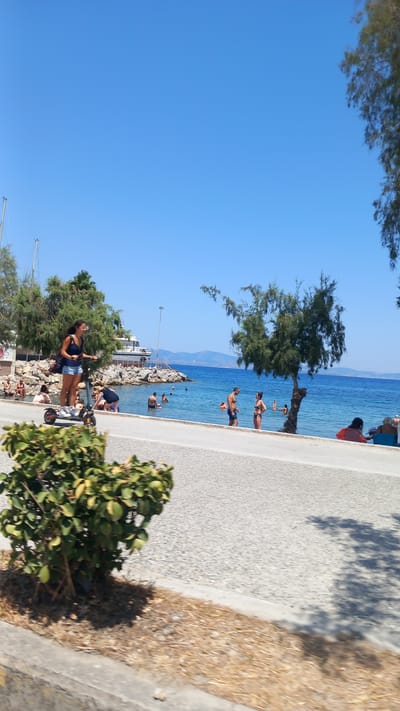 A woman is riding an electric scooter on a paved road next to a beach. She is wearing a dark blue tank top and denim shorts. She has a backpack slung over her left shoulder. In the background, people are swimming and relaxing in the ocean. There are trees and boats visible on the water. The sky is clear and blue. The location is near the city of Heraklion, Crete, Greece, likely at a beach accessible by road. The scene depicts a sunny day at the seaside, with a mix of recreational activities.