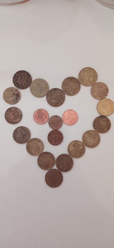 This image displays a collection of Azerbaijani coins arranged on a white surface, forming a loose circular or heart-like shape. The coins are of various denominations, including 5, 10, and 20 qəpik. Some coins are copper-colored, while others appear to be a brass or silver alloy. The visible text on the coins includes "AZƏRBAYCAN RESPUBLİKASI" (Republic of Azerbaijan) and denominations like "10 QƏPİK," "20 QƏPİK," and "5 QƏPİK." Some coins show intricate designs, such as buildings or maps of Azerbaijan. The arrangement of the coins is the primary subject, with no people or animals present. The lighting suggests an indoor setting, likely during the daytime, with no specific weather cues. The overall impression is a still life composition focused on currency.