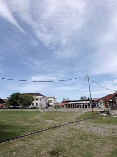 The image depicts an outdoor scene in Mesjid Reuntoh, Indonesia. In the foreground, a large expanse of green grass is visible, with a weathered metal drain cover running horizontally across it. The middle ground features a row of buildings with red-tiled roofs, interspersed with trees and a white multi-story building. Several parked vehicles, including cars and motorcycles, are present in front of the buildings. A green lamppost stands near the center of the image, with power lines extending across the frame. The background is dominated by a blue sky with wispy white clouds. There are no people visible in the image.