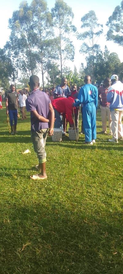 The image shows a group of men standing on a grassy field. The men are gathered around a man in a red shirt who is holding a red blanket. Some of the men are holding sticks, while others are holding baskets. There are several trees in the background. The location is a grassy field in Isukha ICHINA, Kenya, which is located at latitude 0.351 and longitude 34.782. It is a clear day with some clouds in the sky. The men are wearing casual clothes, and the ground is covered in short green grass. The men are all looking in different directions, but they seem to be engaged in conversation. The image appears to have been taken on a smartphone.