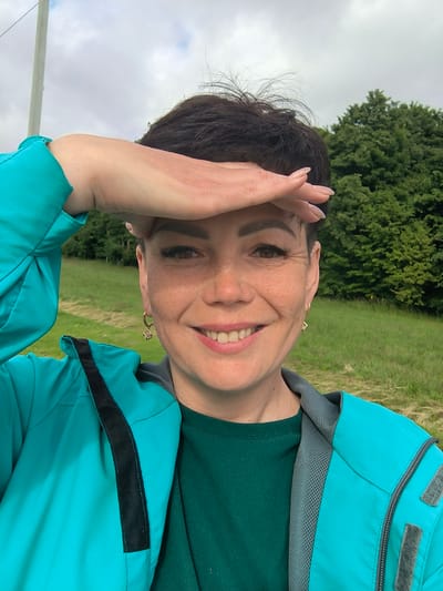 A woman with short, dark hair and freckles is taking a selfie outdoors. She is wearing a teal jacket over a dark green top and has her right hand raised to shield her eyes from the sun. Her expression is a smile. The background shows a grassy field with trees and a cloudy sky. There is a white pole on the left side of the image. The image is not of the city of Laskino, Russia.