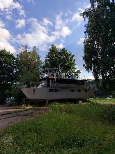 A large, grey vessel, likely a motor yacht or houseboat, is stationary on land, supported by wooden cradles. It is positioned in a clearing surrounded by lush green trees and grass. The sky above is a vibrant blue, dotted with fluffy white clouds. In the background, a body of water, possibly a river or lake, can be seen, along with a distant road and more trees. The scene appears to be in a semi-rural or park-like setting. This scene is located in Birštonas, Lithuania, near the Nemunas River.