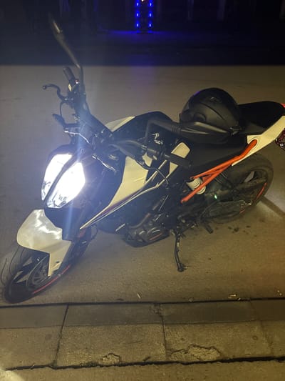 The image shows a parked motorcycle, possibly a KTM, on an urban street in what is likely Mihailovo, Bulgaria. The bike is oriented diagonally in the frame with its headlight illuminated, projecting a bright light. A black motorcycle helmet rests on or near the gas tank. The street is paved, with an adjacent curb visible on the right side. The background is dimly lit and includes a building facade or wall, and a set of blue lights visible to the left. No individuals are visible in the scene.