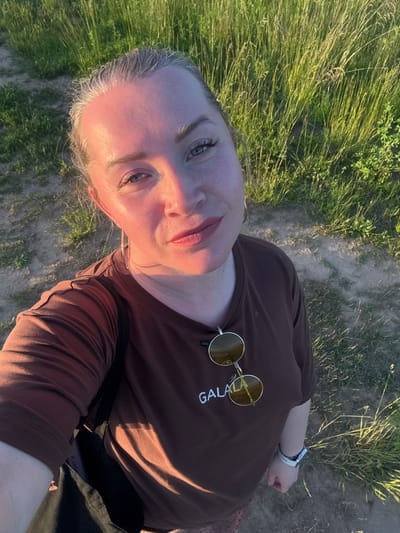 A woman is taking a selfie outdoors, with a field of tall grass and a dirt path in the background. The woman has blonde hair pulled back and is wearing a brown t-shirt with sunglasses hanging from the collar. She is also wearing hoop earrings and a watch on her left wrist. The sun is casting a warm glow on her face. The image appears to have been taken in Sapunov Bor, Russia, based on the context provided.