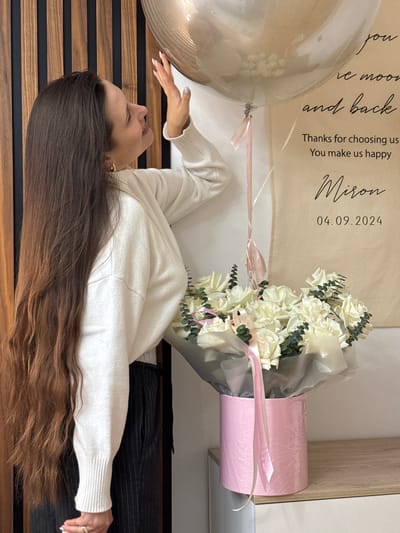 A photograph shows a young woman with long brown hair lying on what appears to be a wooden floor with black accents. She is wearing a white sweater and dark-colored pants. Next to her are a bouquet of white roses in a pink container and a large transparent balloon. Behind the balloon and flowers is a beige sign with the text "Thanks for choosing us. You make us happy Miron 04.09.2024." The setting appears to be indoors. There is no explicit information or detail provided in the image that reveals the specific location in Akulovo (Gp Kubinka), Russia.