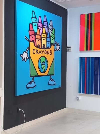 The image shows a square painting on the floor, leaning against a wall. The painting depicts a cartoon-style box of crayons with a world globe on the front. The word "CRAYONS" is printed in black letters on the yellow box. Colorful crayons stick out of the box. Cartoon arms and legs with blue shoes are attached to the box. Above the painting are two horizontally striped paintings: one with pink, red, green, and black stripes; the other with various shades of blue and a red stripe. A keypad is on the wall. A surveillance camera is on the wall. A white cord is on the floor. The floor is covered with a black mat. The location is Port D'andratx, Spain.