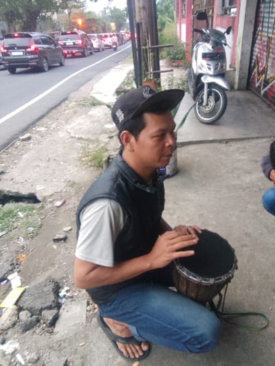 The image depicts a street scene in Cisarua, Indonesia, characterized by a traffic jam. In the foreground, a person wearing a black vest, a light-colored t-shirt, blue jeans, a black baseball cap, and sandals is playing a small drum. The person's hands are positioned on the drumhead, indicating an action of playing. The drum is set on a stand. The ground is paved with concrete and appears somewhat rough, with debris scattered around. In the middle ground, several vehicles are visible, including cars and motorcycles, indicating congested traffic. A scooter is parked on the side of the road. The background shows a building with red and pink accents and some vegetation. The overall lighting suggests it is daytime, possibly late afternoon or early morning.