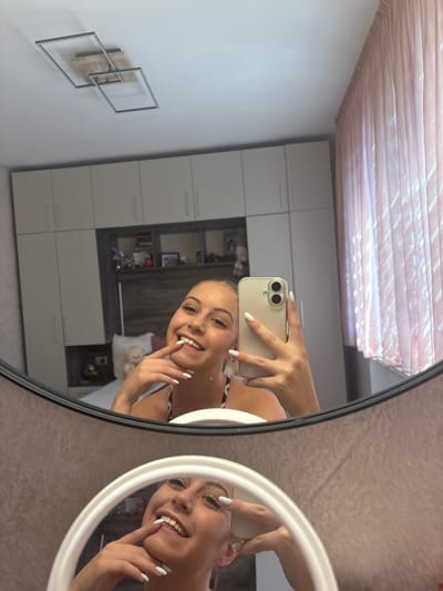 In a bedroom, a young woman is taking a selfie in a large, oval mirror. She is holding a smartphone in her right hand, with her left index finger touching her lips. She is smiling at the camera, and her eyes are bright. The room appears to be well-lit, suggesting it is daytime.
The woman has long, dark hair and is wearing a white tank top. Her nails are painted white. In the reflection, we can see a part of a modern wardrobe with white doors and shelves. There are also some framed pictures on the wall. In the smaller, round mirror on the right, the woman's face is visible again, showing her full smile and playful expression.
The overall atmosphere is casual and cheerful, capturing a moment of self-expression and fun. The setting is a domestic interior, likely a bedroom in Bulgaria, given the location context. There is no visible text in the image.