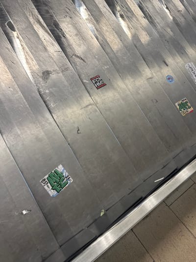 The image displays a close-up, downward-angled view of an unoccupied metallic baggage carousel within an airport terminal, identified as Ben Gurion Airport, Israel. The carousel surface comprises numerous interlocking, brushed metal segments, exhibiting significant wear, scratches, and minor discoloration consistent with heavy use.
Several stickers are affixed to these segments. In the lower-left quadrant, a square sticker features stylized green text against a white and light blue background, with smaller black text visible below it, partially reading "TLVR". In the upper-middle section, a rectangular black sticker with a red border displays the white capital letters "FCK HMS". Further right, a small circular blue sticker is present. Below and to the right of the blue sticker, a multi-colored rectangular sticker with green, white, and red elements features indistinct stylized text. Along the far right edge of the carousel, a white vertical sticker strip displays black Hebrew text, specifically "נתב"ג אילת" (Natbag Eilat), with "Eilat" also visible in English, confirming the location.
The lower-right portion of the frame shows a polished metallic edge trim of the carousel, below which a light-colored, possibly beige, tiled floor is visible. No individuals, luggage, or active operations are depicted.