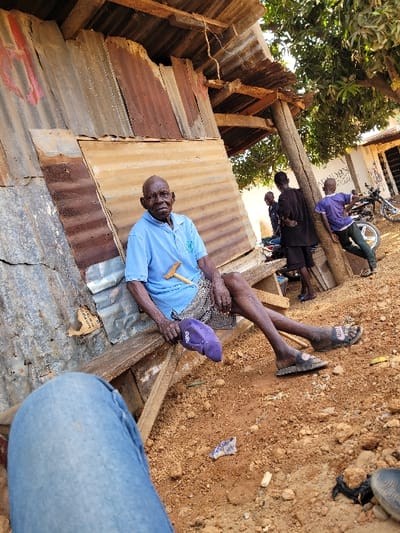 The scene is set outdoors in Ube, Nigeria. An elderly, bald man with dark skin sits on a simple wooden bench, leaning against a wall constructed from various corrugated metal sheets, some rusted. He wears a light blue polo shirt, patterned shorts, and dark sandals. His right hand holds a light-colored wooden object, and a purple cap rests on the bench near his left leg. His legs are extended, with his feet resting on the reddish-brown, dusty ground, which is sparsely littered with small debris, including a white and blue wrapper.
Behind and to the right of the seated man, a wooden pole supports a corrugated metal overhang. Three other individuals are visible in the background: two men in dark shirts stand, partially obscured, and a third man in a purple shirt and light trousers leans against the wooden pole, facing away from the camera. Further in the background, a light-colored building with dark windows or doorways is visible, and a motorcycle is parked nearby. A large tree with green leaves occupies the upper right portion of the frame, casting shadows. The image is captured from a low vantage point, with the photographer's blue-jeaned legs visible in the bottom left foreground.