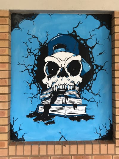 A vertical mural is displayed within a rectangular, brick-framed recess in a structure located in Midrand, South Africa. The artwork features a stylized white human skull with prominent black eye sockets and a jagged grin, wearing a blue baseball cap with the bill positioned backward. The skull is centrally located above a stack of approximately five white books with blue spine details. A black, viscous liquid drips from the skull's mouth, flowing over and between the books, creating pools beneath them. The background of the mural is a solid light blue, from which black, gnarled, root-like or crack-like patterns emanate, primarily from behind the skull, extending towards the edges of the panel. The signatures "M. MALEMA" and "T.M. MALEMA" are visible in black text in the lower right quadrant of the artwork. The mural is framed by light brown brickwork on its left and bottom sides, with a thin white trim bordering its upper edge.