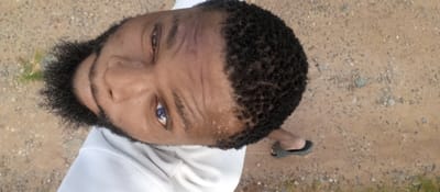 A close-up, high-angle shot features a dark-skinned man with short, curly hair and a full black beard. He is wearing a white top. His eyes are a striking blue color. The background appears to be a dirt or gravel surface, typical of an unpaved road or ground area. A portion of a black sandal or shoe is visible in the lower right corner. Given the location of Magboro, Nigeria, it is probable that the ground is sandy or laterite soil.