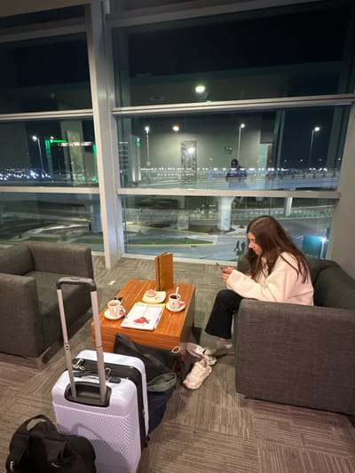 The image depicts an indoor airport lounge or waiting area located in San Miguel Jaltocan, Mexico, at night. A lone female individual with long brown hair is seated in a dark gray armchair, wearing a light-colored top and dark pants, actively engaged with a smartphone held in both hands. Her pink sneakers are placed on the patterned carpet beside her feet. Adjacent to her and centered in the frame is a wooden coffee table. On the table are two white ceramic cups with saucers (containing dark beverages), a small plate holding a pastry, a brown stand-up menu, and a magazine titled "BAJA FLAVORS." To the left of the table, a light lavender hard-shell rolling suitcase with an extended handle stands on the carpet, accompanied by two dark-colored soft bags. Large windows behind the seating area provide a panoramic view of an illuminated airport environment at night, featuring a multi-lane elevated roadway, various distant structures including one with a green "Holiday Inn Express" sign, and numerous artificial lights across the landscape.