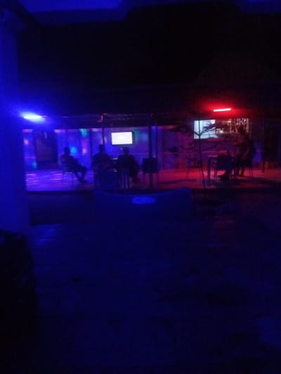 This is a nighttime photograph taken at the Blue Sea Bar and Restaurant in Sabon Gari, Nassarawa, Nigeria. The scene is illuminated by a combination of blue and red lights. In the background, several patrons are seated at tables, and a television screen is visible. Some of the patrons appear to be engaged in conversation, while others are observing the screen. The establishment has an open-air or semi-open-air seating area, with a roof structure visible in the upper portion of the image. The overall atmosphere suggests a lively social gathering or entertainment venue.