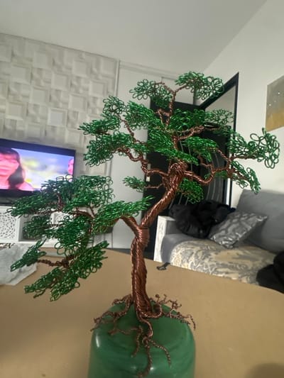 The image depicts a wire-sculpted bonsai tree, serving as the primary subject, positioned centrally on a light brown, flat surface. The tree's trunk and branches are formed from twisted copper-colored wire, while the foliage is intricately crafted from green wire. The sculpture is mounted on a smooth, rounded green base.

In the background, to the left, a flat-screen television is visible, displaying the face of a woman. Below the television is a white console table or cabinet with a geometric, possibly carved, open-work pattern. The wall behind this area features a raised, textured pattern of interconnected squares. To the right of the bonsai tree, a segment of a light-colored sofa with a patterned upholstery is visible, adorned with a dark pillow and a metallic-sheen, patterned grey pillow. A dark, undefined object rests on the sofa. A dark vertical structure, likely a doorway or partition edge, is present further right, with a framed piece of abstract art partially visible on the wall beyond. The scene is illuminated by ambient indoor lighting. This indoor environment is situated in Marcoussis, France.