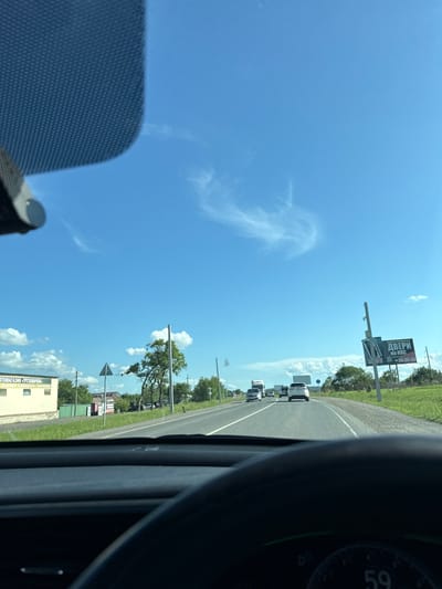 The image shows a view from the interior of a car traveling on a highway in Timiryazevskiy, Russia. The sky is bright blue with wispy clouds. On the left side of the road, there is a light-colored building with the text "АВТОМАГАЗИН - ГРУЗОВИЧОК" which translates to "Car Shop - Trucker". A green fence is behind the building. Further down the road, there is a billboard on the right advertising "ДВЕРИ НА КПС" which translates to "Doors to KPS". Several vehicles, including cars and a truck, are on the highway. The dashboard of the car is visible in the foreground, with a speedometer displaying "59". The environment appears to be a rural or suburban setting with trees and green grass lining the road.