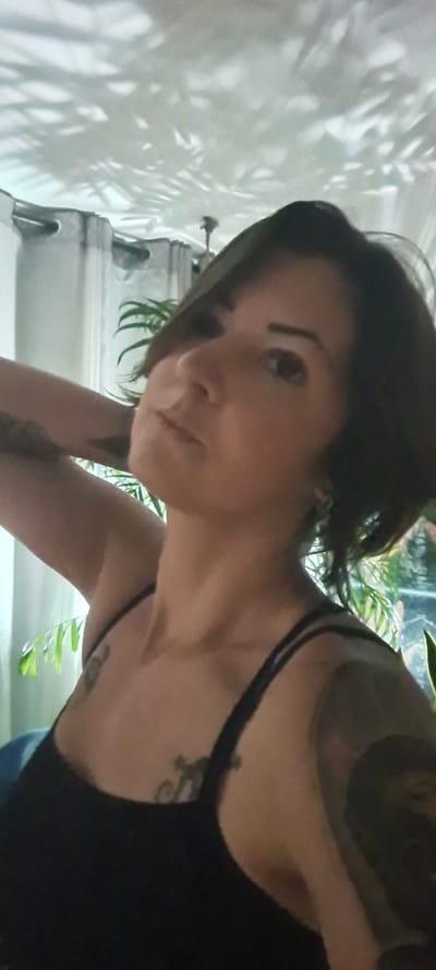 A woman with short brown hair, fair skin, and tattoos is seen from a low angle. She is wearing a black tank top. The background includes a white wall with the shadow of a plant, and glimpses of greenery suggesting indoor plants. While the city of Nahariyya, Israel is mentioned, there is no visual indication of the location being Nahariyya or any other specific city.