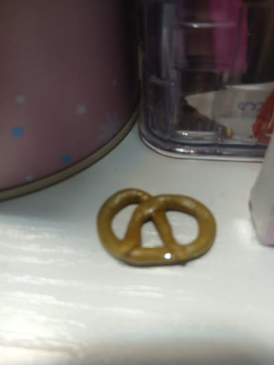 The media file is a close-up, low-quality image of a pretzel. The pretzel is golden brown and appears to be a classic knot shape. It is placed on a white surface, possibly a countertop or table. To the left of the pretzel, there is a blurry container with some items inside, including what appears to be pink plastic and a white wrapper with some text on it. Below the container and to the left of the pretzel, a portion of a pink, round object with blue polka dots and star-like shapes is visible, possibly a bowl or a mat. The lighting in the image is somewhat harsh and creates reflections on the white surface and the pretzel. No specific time of day, weather, or emotional cues can be determined due to the low quality and the absence of context. There is no discernible text from the image, except for what may be on the blurry wrapper. The scene appears to be indoors, in a domestic setting.