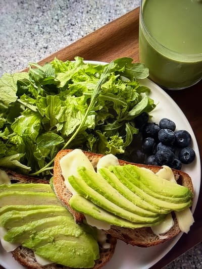 The image displays a close-up, overhead view of a healthy meal set indoors, likely in Ros Na Rí, Ireland. On the left, a clear glass filled with a vibrant light green smoothie or juice rests on a wooden surface. A white, oval-shaped plate dominates the center, holding various food items.

On the plate, a large serving of fresh green salad, appearing to be a mix of lettuce and arugula, occupies the lower left section. Adjacent to the salad, a small pile of dark blue blueberries adds a fruity element. The primary components of the meal are two slices of what appears to be whole-grain toast. Each toast slice is generously topped with creamy white cheese, possibly melted, and meticulously fanned slices of bright green avocado.

The plate and glass are positioned on a warm-toned wooden board or tray, which itself is resting on a light-colored, speckled countertop or table. The lighting suggests daytime, enhancing the natural colors of the fresh ingredients. There are no people visible, nor any discernible text in the frame. The overall scene conveys a focus on fresh, nutritious eating with a clean and appealing presentation.