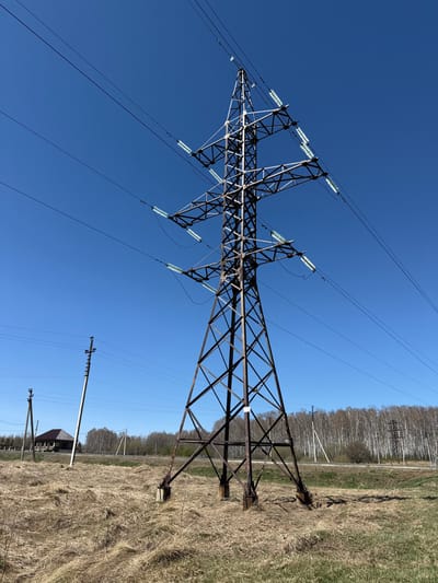 A large, metal electrical tower, supporting multiple power lines, stands in a field of dry grass. It is located in Verkh-Chebula, Russia, at the coordinates 56.019, 87.587. The tower is rusty and weathered, and the lines are strung between insulators. Two smaller power poles are visible in the background, along with a small, wooden structure to the left of the tower. A line of birch trees is visible in the distance. The sky is clear and blue.