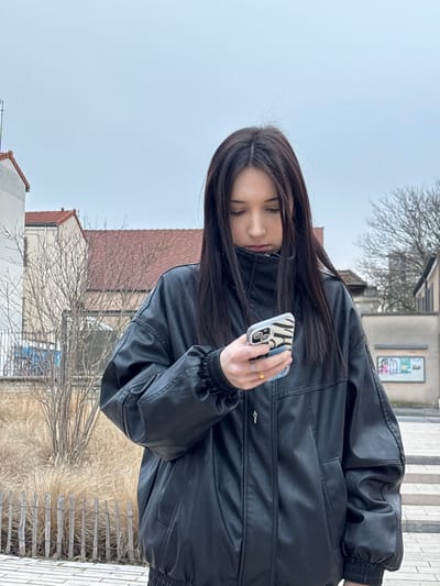 A woman wearing a black leather jacket is looking down at her phone, which has a zebra-striped case. She is standing outside near some buildings and dry, tall grass. The sky is overcast. Her dark hair hangs straight down. She is wearing a couple of rings. The scene appears to be a casual, everyday moment captured in a photograph.