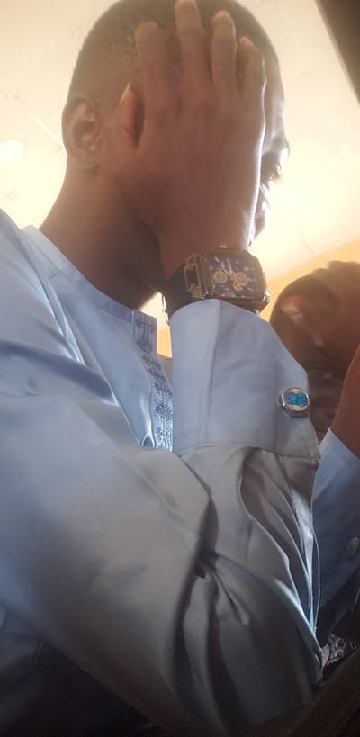 The image shows a close-up of a man's head and upper body, seen from a low angle. His right hand is covering his face. He is wearing a light blue collared shirt with patterned trim on the placket and a watch on his left wrist. A cufflink with a blue stone is visible on his left shirt cuff. The background is out of focus, but appears to be an indoor setting with a light-colored ceiling. Another person's head is visible in the background on the right side of the image. The overall impression is that the man is either shielding himself or expressing distress.