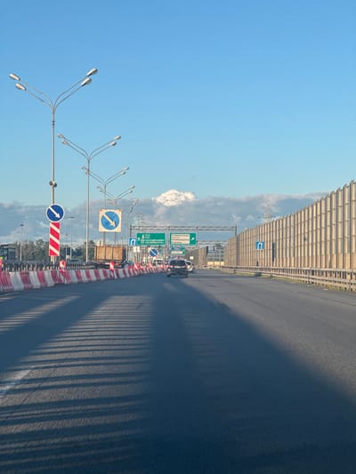 The image depicts a highway scene in Lisy Nos, Russia, under a bright blue sky with scattered clouds. The perspective is from inside a vehicle, looking forward. A roadway stretches into the distance, bordered on the right by a tall, beige sound barrier. On the left, a row of red and white traffic cones and barriers delineates a construction or lane closure area. Several streetlights with multiple lamps are visible overhead, along with various road signs. One sign is a blue circle with a white arrow pointing diagonally down and to the right, indicating a mandatory turn. Another sign, also blue with a white arrow, indicates a change in lane direction. In the distance, green highway signs display destinations including "Primorskoye Highway." A few vehicles, including a black SUV and a white car, are on the road. The sun casts long, striped shadows across the asphalt.