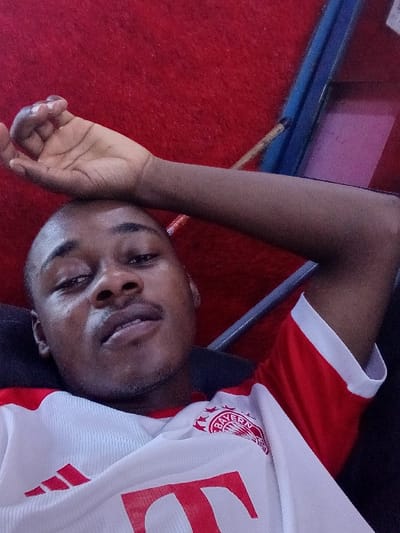 An adult male with dark complexion, short dark hair, and light stubble on his chin and upper lip is depicted in a close-up, upward-angled shot. He is wearing a white and red Bayern Munich football club jersey, identifiable by the club crest on the left chest and a partial "T" logo in red on the front. His left arm is bent, with his hand resting near his forehead, while his gaze is directed towards the camera. The subject appears to be lying on his back, supported by a dark headrest or cushion. The background consists primarily of a textured red surface, resembling carpet or upholstered material. A blue-colored linear object, possibly a frame or rail, is visible in the upper right quadrant of the image, alongside a blurred object with a magenta hue. The specific location within the city of Dorayi, Nigeria, is not visually discernible from the image content.