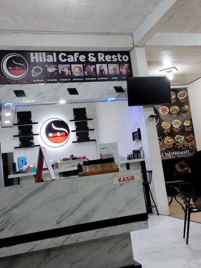 The image shows the interior of Hilal Cafe & Resto in Blangkarimueng, Indonesia. The cafe features a modern design with a marble-patterned counter and shelves. A prominent sign above the counter displays the cafe's name and images of various beverages. A menu board is visible on the right wall, showcasing food items. In the foreground, a "KASIR" (cashier) sign is displayed on the counter. A man is seated at a table in the background, appearing to be a customer. The overall environment suggests a casual dining and cafe setting.