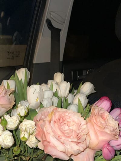 The image displays a large floral bouquet positioned within the passenger compartment of a vehicle at night, reportedly in the city of Базарово, Russia. The bouquet primarily features light pink garden roses, numerous white tulips with prominent green stems, smaller white cluster roses or buds, and a few pink tulips. Some of the larger pink roses exhibit minor yellowing on their petal edges. The background reveals elements of the car's interior: a dark window reflecting a light-colored box with black symbols, and a black seatbelt extending vertically from a light grey interior panel. The scene is illuminated by an unseen light source, contrasting with the dark exterior visible through the window. No individuals or specific actions are depicted.