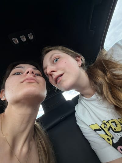 The image shows two women looking at the camera, appearing to be in a vehicle. The woman at the top of the image has lighter hair and is wearing a white t-shirt. The woman at the bottom has darker hair and is wearing a delicate necklace. The interior of the vehicle is visible, showing a black ceiling and seat. The location is not discernible from the image.