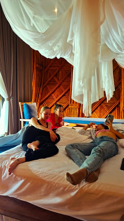 Three young adult females are seen relaxing on a large bed in a bedroom located in Mù Cang Chải, Vietnam. The room features a prominent wall constructed from interwoven bamboo, forming a lattice pattern. A white, sheer canopy hangs over the bed.

On the left, a light-haired female, wearing a black tank top and black pants, is semi-reclined, looking down at a smartphone in her hands. Her bare feet are visible, and a small tattoo is on the top of her left foot. Adjacent to her, in the middle, a dark-haired female in a red top with a distinct collar sits upright, observing something to her right. A small dish is positioned near her. On the far right, a female with medium-length hair is lying on her back, wearing a pink crop top, light blue ripped jeans, and light brown polka-dotted socks. She holds a smartphone with a visible camera array above her head, engaging with the screen. A dark object, possibly another phone, rests on the bed near her feet.

The room is brightly lit, with light entering from a window on the far left, where drapes are pulled back. A wooden chair with a blue cushion is partially visible on the left. The bed is covered with white sheets and pillows.