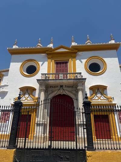 The image shows the exterior of the Plaza de Toros de la Maestranza in Seville, Spain, on a clear, sunny day. The building is predominantly white with yellow accents and features a grand entrance with red double doors. Above the entrance, a balcony overlooks the plaza, and the building is adorned with circular windows, decorative elements, and ornate finials on the roof. A black wrought-iron fence with spikes and decorative panels encloses the entrance. The fence is set against a yellow brick wall. The sky is a vibrant blue. There are no people visible in the image.