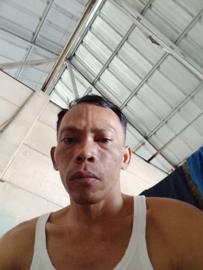 A head-and-shoulders selfie of a man is shown, taken from a low angle. The man has dark hair, brown eyes, and a light complexion. He is wearing a white tank top. The background consists of a corrugated metal roof with visible structural supports and a white wall to the left. A dark blue fabric is visible to the right of the man. The scene appears to be indoors, possibly in a workshop or covered outdoor area. The image was taken in Bukit Ketok, Indonesia.